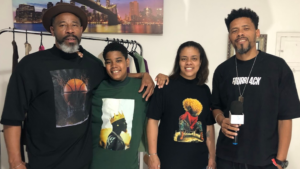 Quatro pessoas sorrindo para a câmera, vestindo camisetas com estampas de moda streetwear. Um homem com chapéu, um jovem, uma mulher e um homem segurando um microfone.