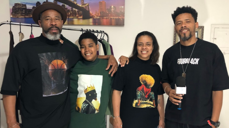 Quatro pessoas sorrindo para a câmera, vestindo camisetas com estampas de moda streetwear. Um homem com chapéu, um jovem, uma mulher e um homem segurando um microfone.