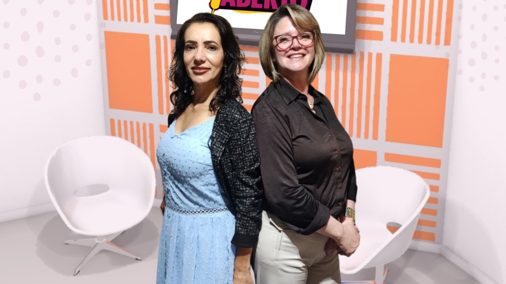 Duas mulheres Marcia Perinoti e Lia Belli Custodio, uma de vestido azul e outra de blusa escura, posando de costas uma para a outra em um estúdio de TV com cadeiras brancas e um painel laranja ao fundo.