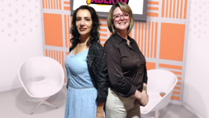 Duas mulheres Marcia Perinoti e Lia Belli Custodio, uma de vestido azul e outra de blusa escura, posando de costas uma para a outra em um estúdio de TV com cadeiras brancas e um painel laranja ao fundo.