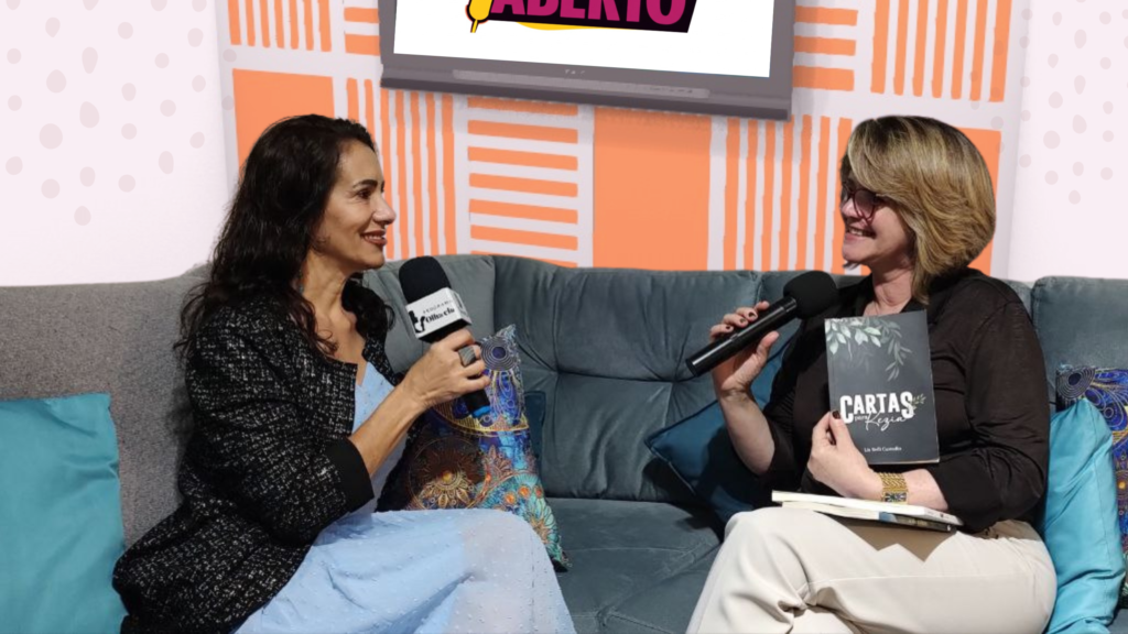 Marcia Perinoti, à esquerda, entrevista a escritora Lia Belli Custodio, à direita. As duas estão sentadas em um sofá em um estúdio, e Lia segura seu livro "Cartas Falsas".