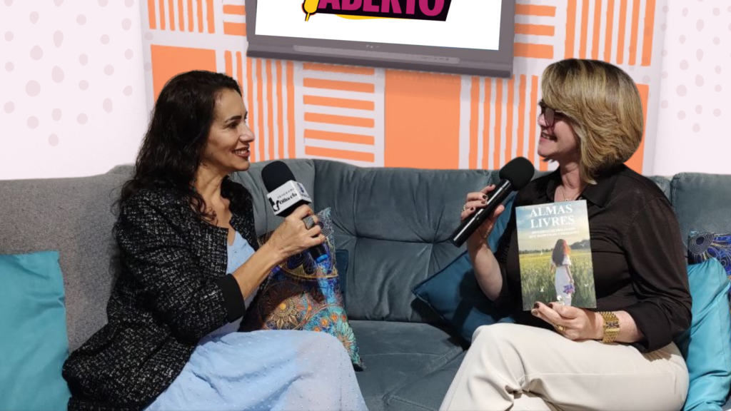 Marcia Perinoti, à esquerda, entrevista a escritora Lia Belli Custodio, à direita. As duas estão sentadas em um sofá em um estúdio, e Lia segura seu livro "Almas Livres".