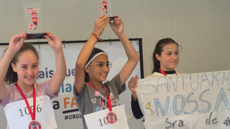 Três meninas coroinhas no pódio da 8ª Corrida de São Tarcísio, sorrindo e exibindo seus troféus; Maria Luiza está ao centro segurando o troféu de 1º lugar.
