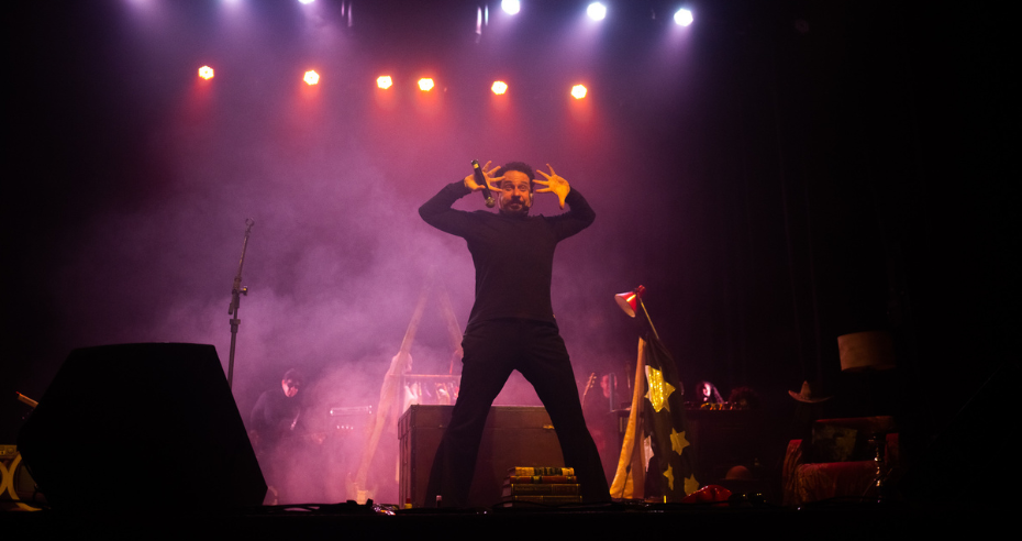 Ator em cena interpreta Raul Seixas com expressão intensa, sob iluminação teatral e fumaça, durante o espetáculo “Raul Seixas, O Musical”.